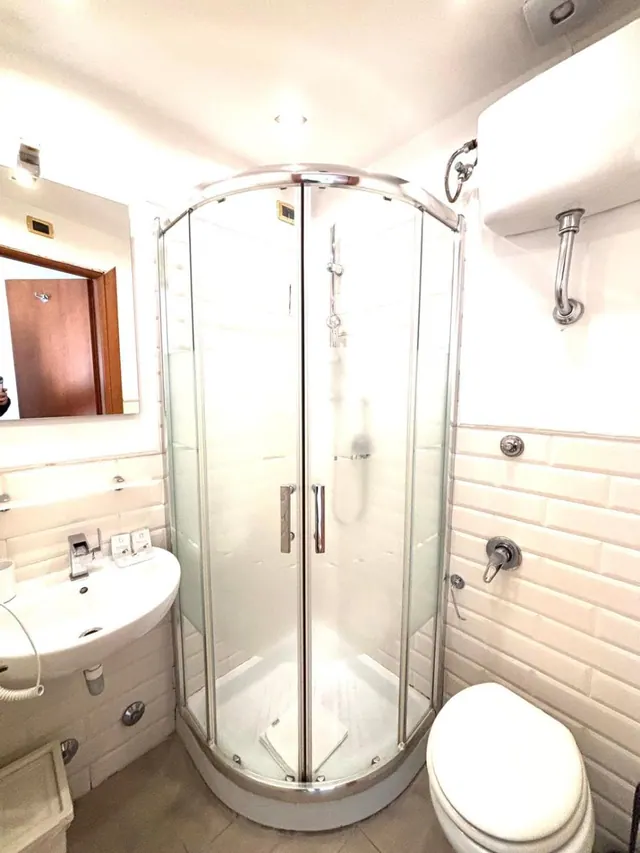A bathroom with a curved glass corner shower, a wall-mounted sink with a mirror above it, and a toilet beside white tiled walls.