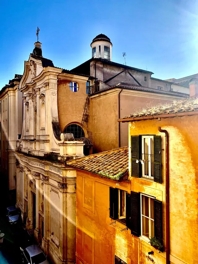 A view from a window over neighbouring rooftops, a baroque church facade, a street with parked cars, and a yellow building under a clear blue sky.