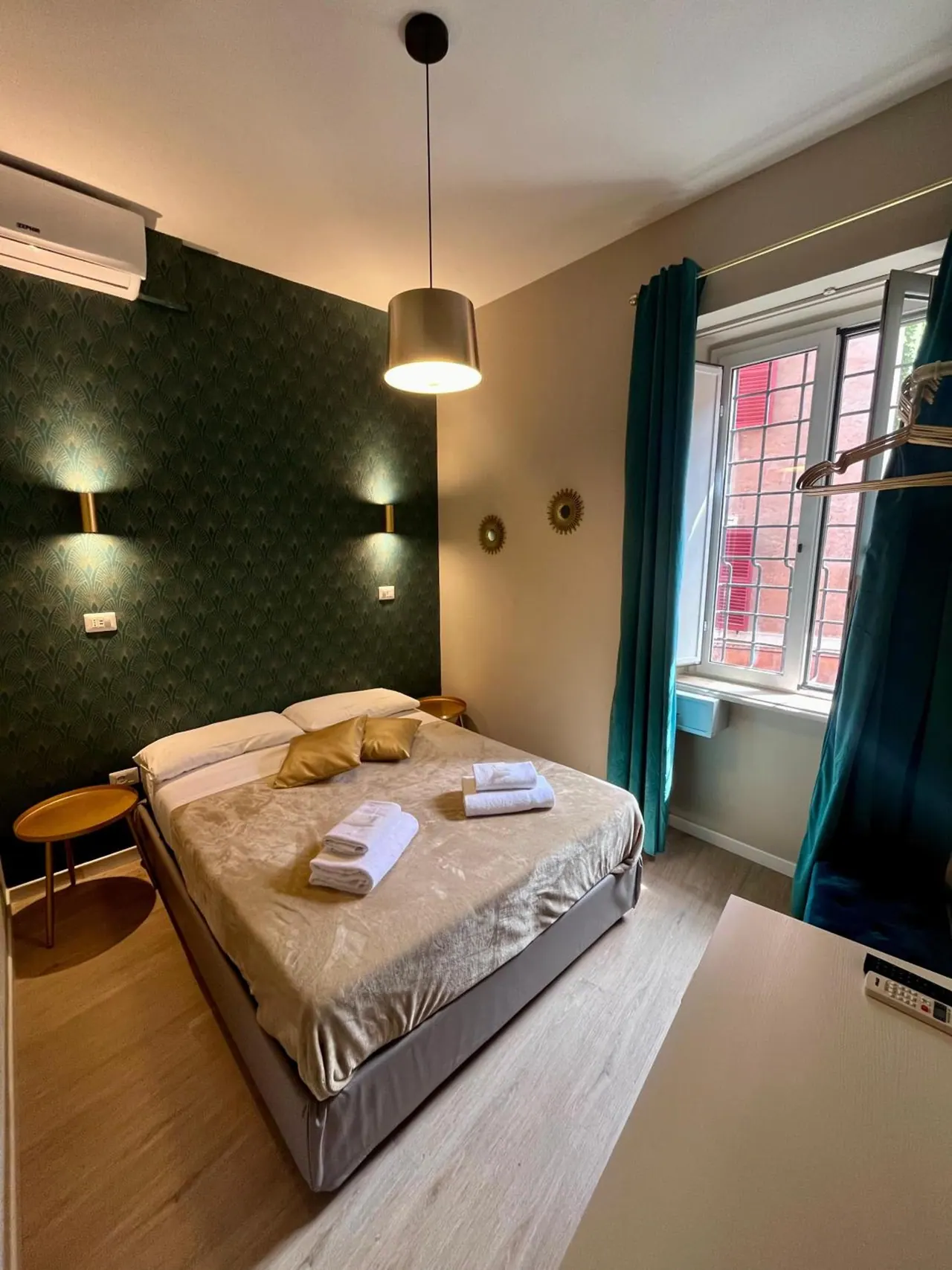 A double bed with folded towels on it sits in a bedroom with green patterned wallpaper, a wall-mounted air conditioner and a window facing a brick building.