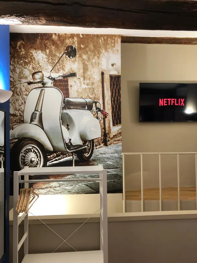 A room with a wall mural of a vintage scooter, a wall-mounted television showing Netflix, a white clothes rack with wooden hangers, and a white railing above a wooden floor.