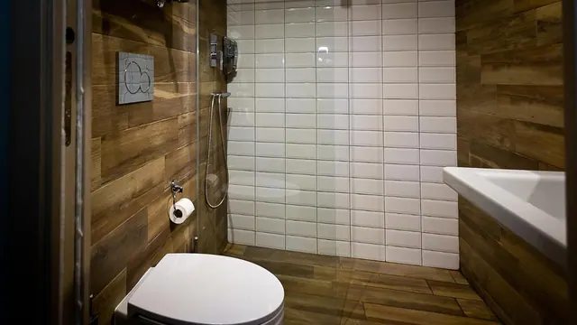 A bathroom with wood-effect tiled walls, a wall-mounted toilet, and a walk-in shower with white subway tiles behind a glass screen, with a white basin on the right.