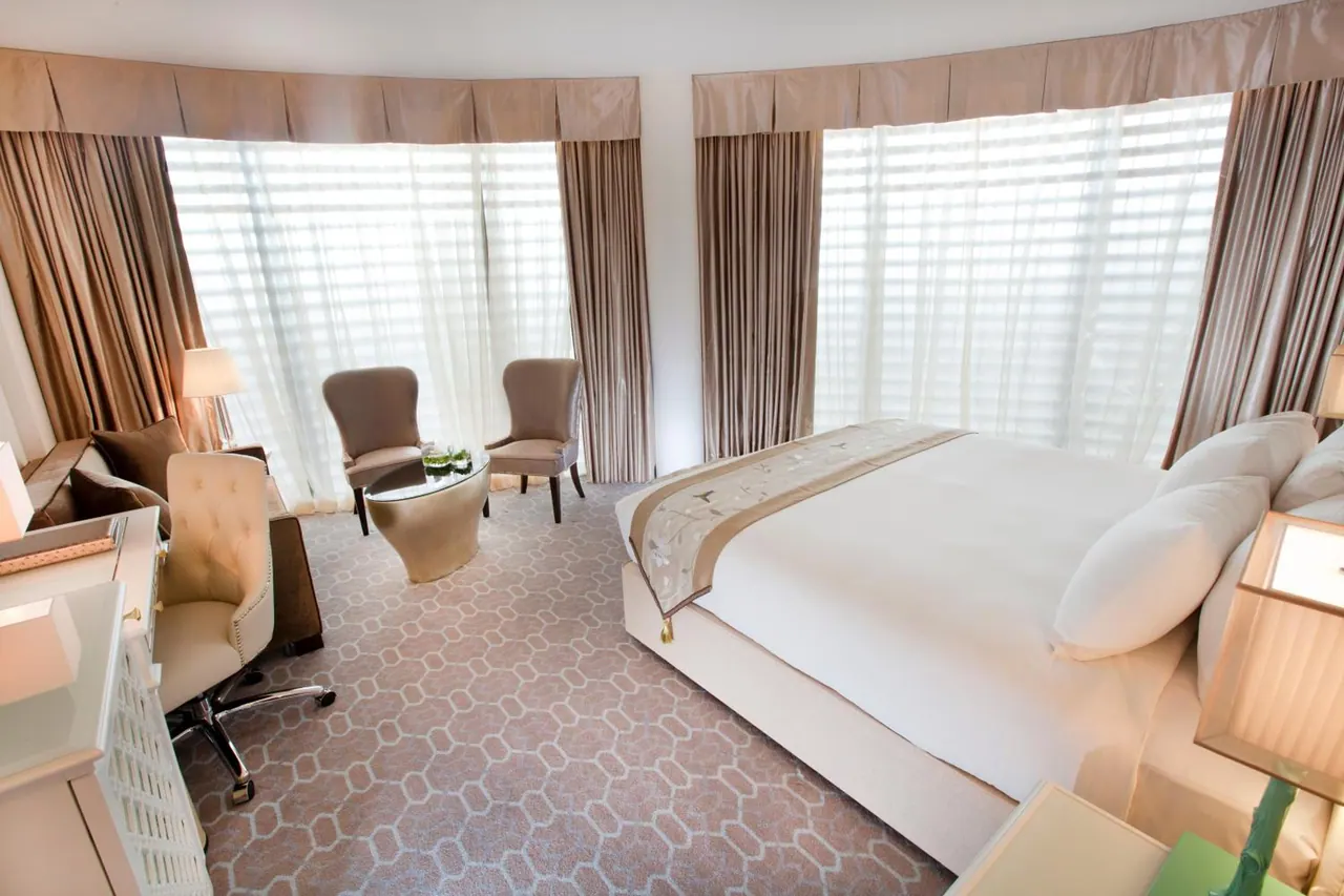A bedroom with a large bed, two armchairs beside a small round table, a sofa and a desk with chair, and tall curved windows covered by sheer blinds.