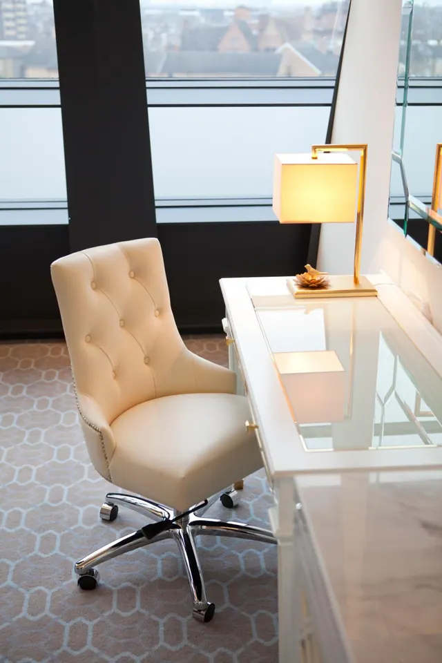 A white tufted leather desk chair and glass-topped desk with a gold lamp stand beside a window looking out over city rooftops.
