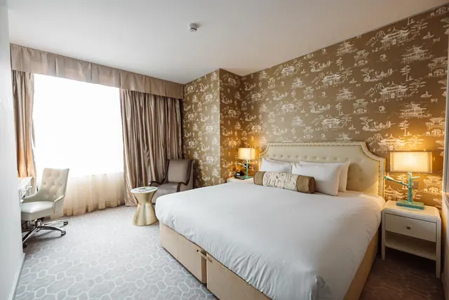 A hotel bedroom with a large white king bed, patterned wallpaper, two bedside lamps, an armchair, and a white desk chair by a bright window.