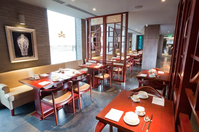 A restaurant dining room with red wooden tables and chairs, cream banquette seating, carved red screen dividers and a large window on the left.
