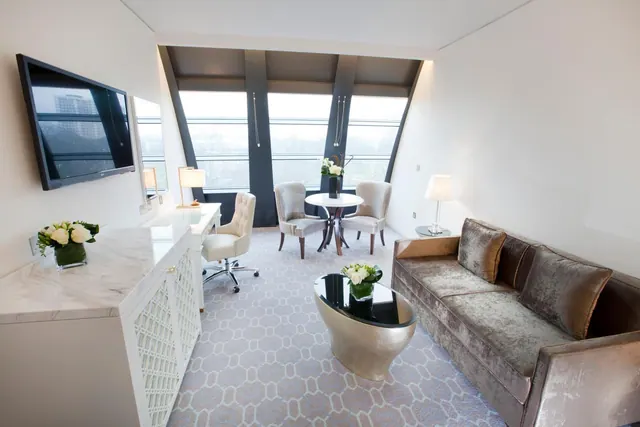 A hotel suite living area with a grey velvet sofa, wall-mounted television, white writing desk and chair, and tall windows looking out over buildings and trees.