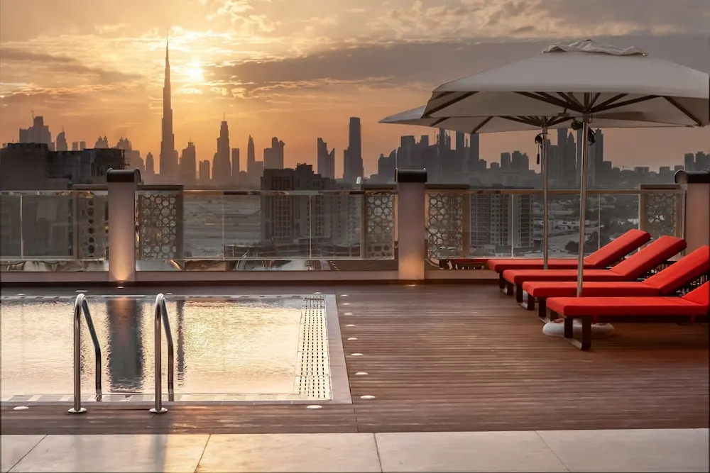 A small rooftop pool with red sun loungers and umbrellas overlooks a city skyline with the Burj Khalifa at sunset.