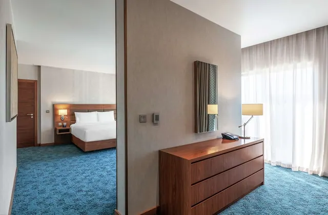 A hotel suite with a wooden dresser, a table lamp and a telephone in the foreground, with a bed visible through a doorway on the left and sheer curtains over a window on the right.