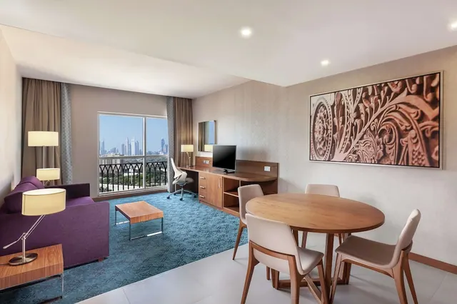A furnished living area with a purple sofa, a round wooden dining table with four chairs, a desk with a television, and a window showing a city skyline.