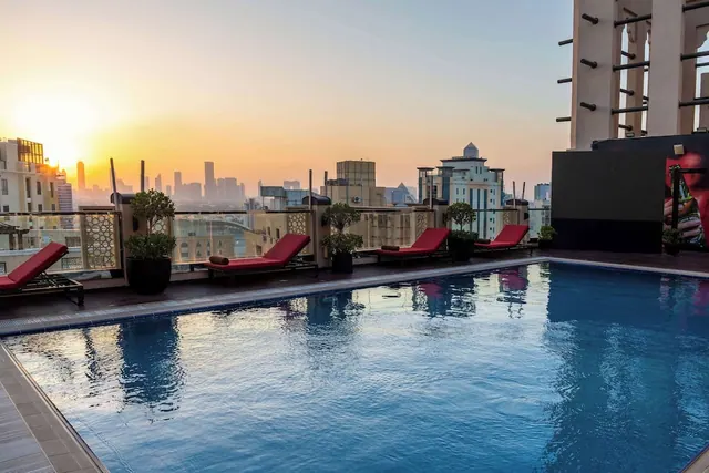 A rooftop swimming pool with red sun loungers and potted plants overlooks a city skyline at sunset.
