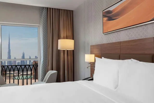 A bedroom with a made king bed, a floor lamp and an armchair, with a balcony door showing the Burj Khalifa and city skyline.