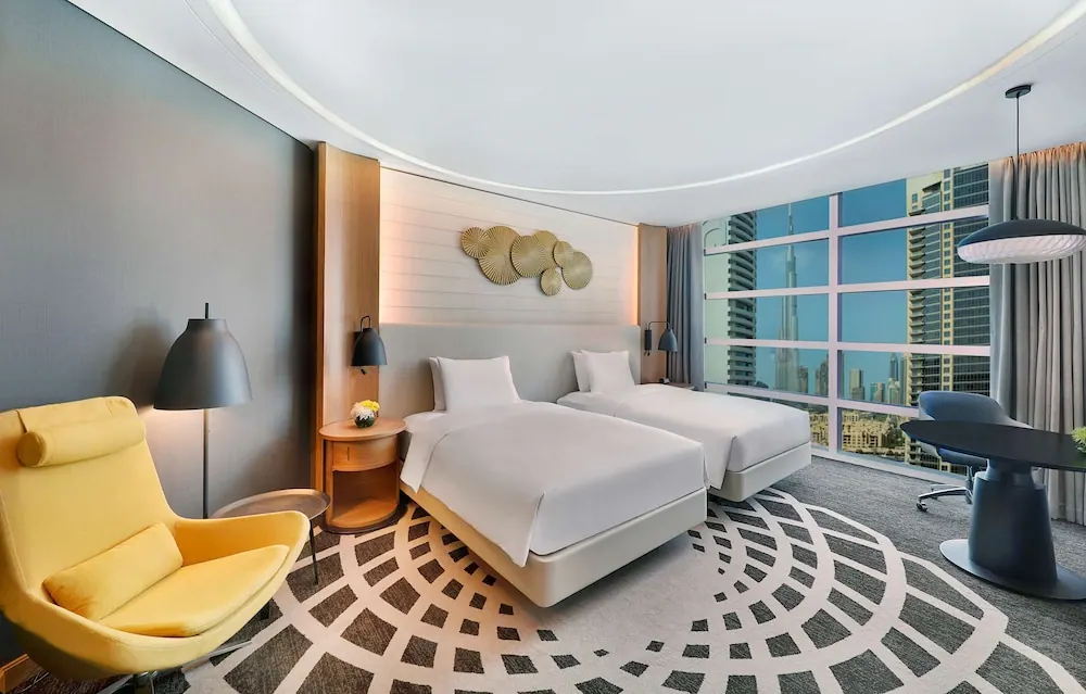 A twin-bedded hotel room with a yellow armchair, a desk, and floor-to-ceiling windows showing the Burj Khalifa and surrounding towers.