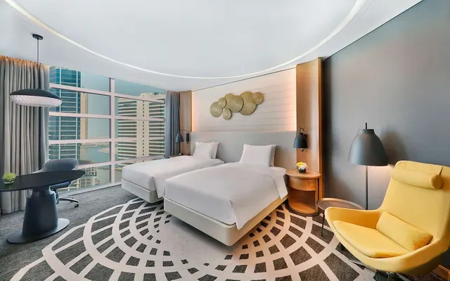 A hotel bedroom with two single beds, a desk and chair, a yellow armchair, and floor-to-ceiling windows overlooking high-rise buildings and water.