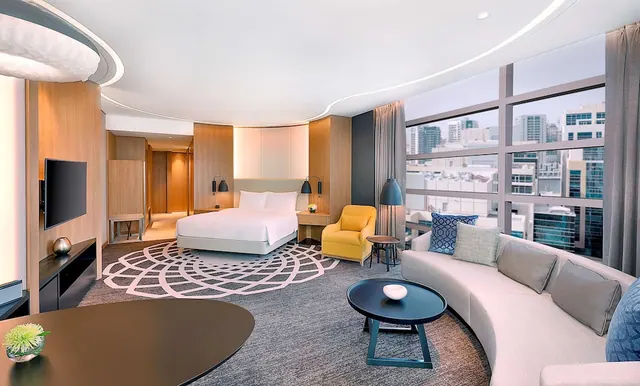 A suite with a made bed, a curved sofa, a yellow armchair, a wall-mounted television and floor-to-ceiling windows looking out over city buildings.