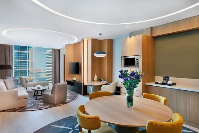 A staged suite living and dining area with a round dining table, lounge seating, a wall-mounted TV and microwave, and floor-to-ceiling windows showing towers and water.