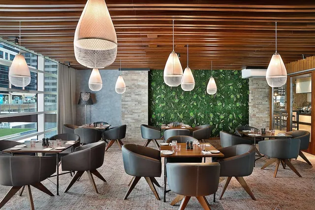 A restaurant dining room with grey leather swivel chairs, wooden tables set for dining, pendant lights, a slatted wood ceiling, a green living wall and floor-to-ceiling windows showing high-rise buildings.
