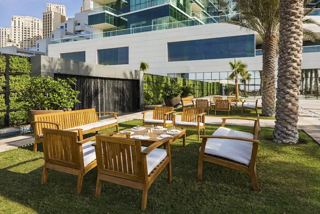 Teak lounge chairs and a low table are set on a lawn beside a water wall and green wall, with palm trees and a glass hotel building behind.