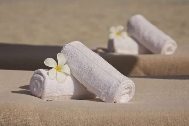 Rolled white towels, two of them topped with white frangipani flowers, rest on tan sun lounger cushions in bright sunlight.