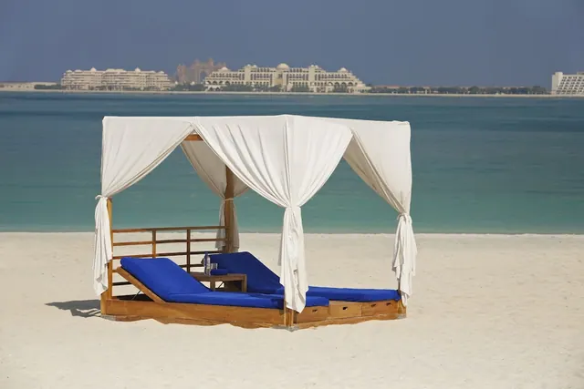 A white-curtained canopy daybed with two blue cushioned loungers stands on a sandy beach, facing the sea and hotel buildings across the water.