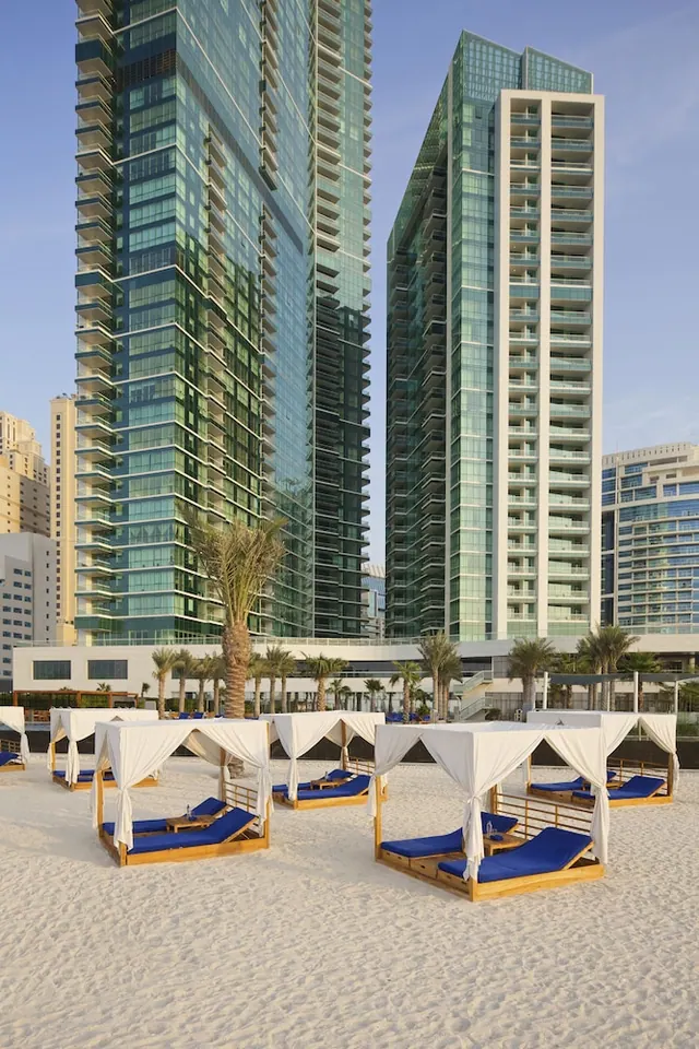 White-canopied cabana beds with blue cushions stand on a sandy beach, with tall glass residential towers and palm trees behind.