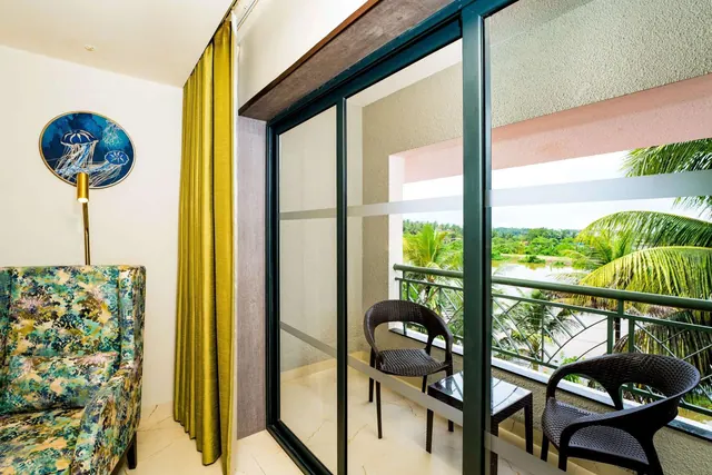 A furnished balcony overlooks lush tropical greenery from a bright room with a patterned armchair.