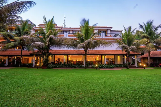 A tropical hotel building with tiled roofs and balconies stands behind a broad manicured lawn and palm trees.
