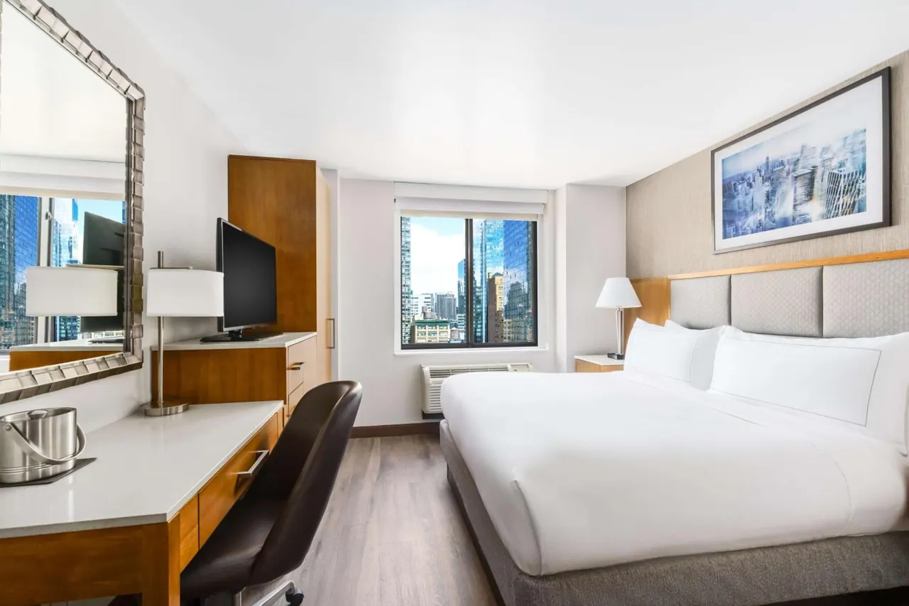 A bedroom with a king bed, a wooden desk with a mirror, a television, and a window looking out over city skyscrapers.