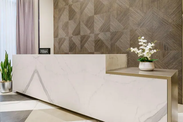 A lobby reception desk with a marble front and a patterned wood-panel wall stands beside sheer and lilac curtains, with a white orchid arrangement on a ledge and a Hilton Honors sign on the wall.