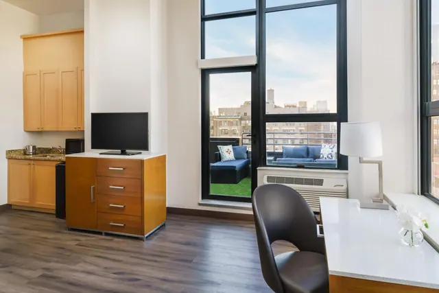 A hotel room with a kitchenette, wall-mounted television, wood cabinet and desk, and a glass door opening onto a balcony with a blue sofa and apartment buildings beyond.