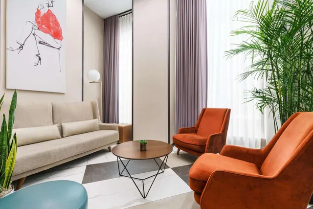 A lounge seating area with a beige sofa, two orange armchairs, a round wooden coffee table, potted palms and a framed fashion sketch on the wall.