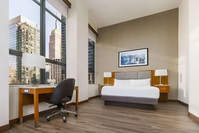 A bedroom with a double bed, a wooden work desk with an office chair, and floor-to-ceiling windows looking out at the Empire State Building.