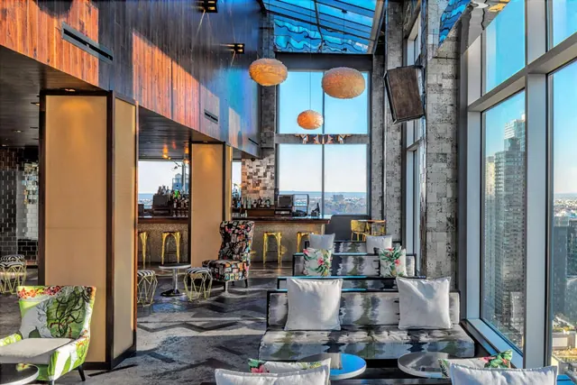 A bar and lounge area with sofas, armchairs, pendant lamps and floor-to-ceiling windows overlooking city buildings.