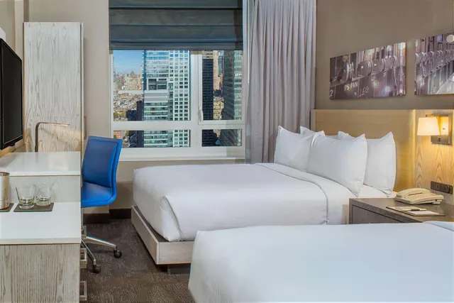 A hotel bedroom with two double beds, a desk with a blue chair, a wall-mounted television, and a window looking out over city skyscrapers.