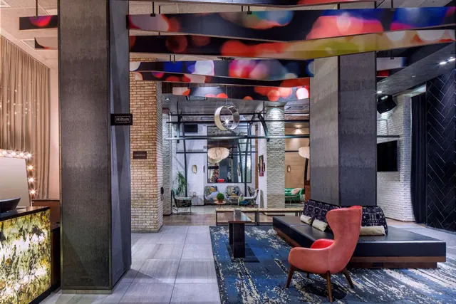 A hotel lobby with a coral armchair and dark leather bench on a blue patterned rug, colorful ceiling panels overhead, and a reception desk at left.