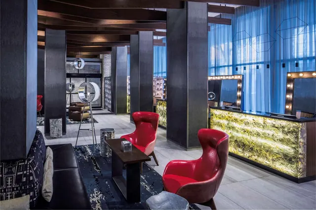 A hotel lobby with red leather armchairs, a dark leather sofa, and a reception desk with a backlit stone front and illuminated vanity mirrors.