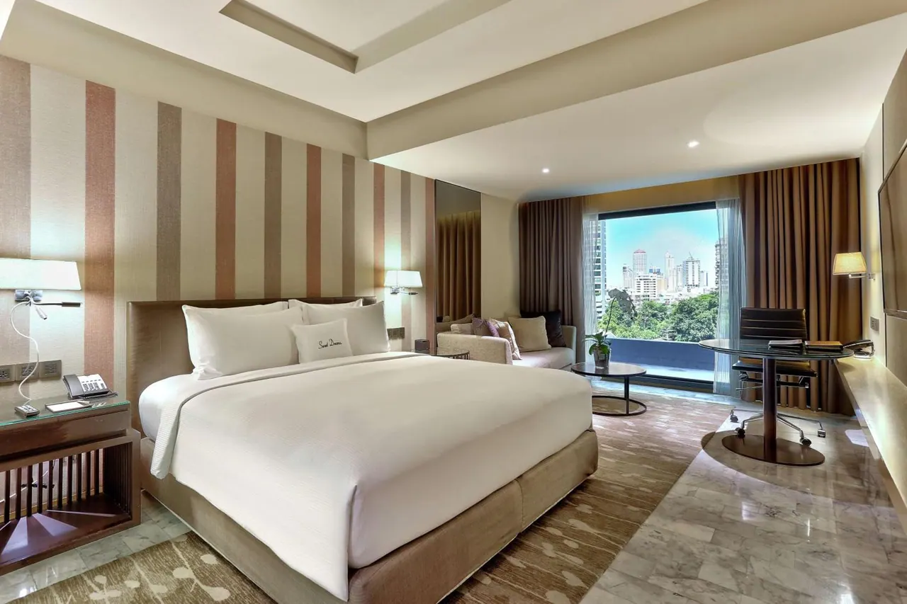 A king bed with a striped feature wall, a seating area, a work desk and a large window overlooking city high-rises and trees.