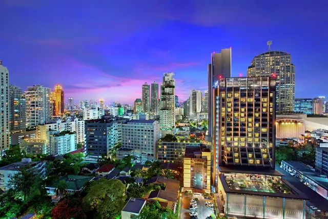 Dusk view over a Bangkok skyline with illuminated high-rise towers and a lit hotel building with a parking area in the foreground.