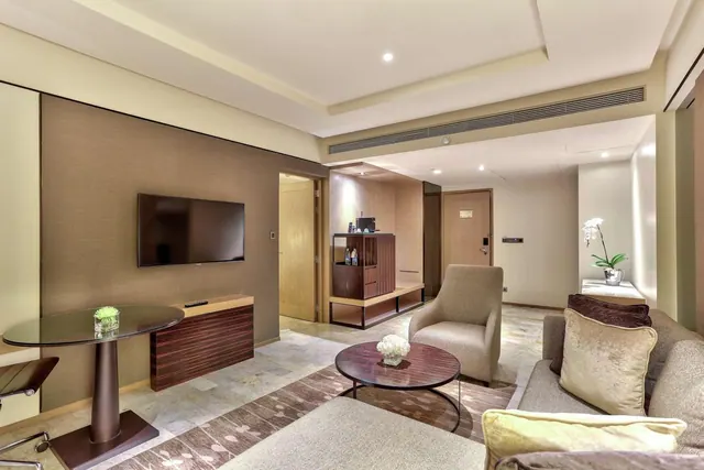 A hotel suite living area with a wall-mounted television, a round glass table with a chair, a wooden sideboard, an armchair, a sofa with cushions and a round coffee table holding a white flower arrangement.
