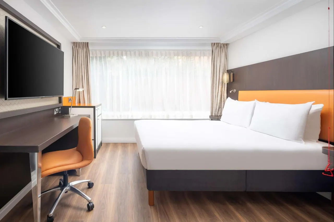 A double bed with white bedding and an orange headboard stands beside a wooden desk, orange chair and wall-mounted television in a hotel bedroom.