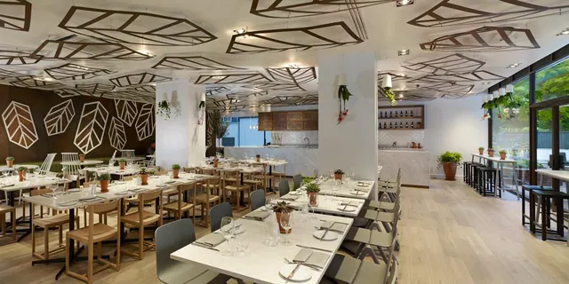 A restaurant dining room with set tables and wooden and grey chairs beneath a leaf-patterned ceiling, with a bar at the back and windows on the right showing greenery outside.