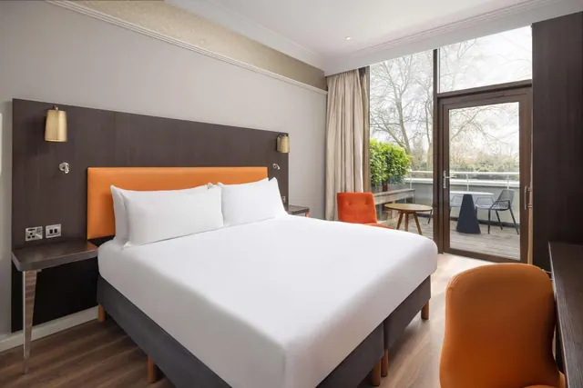 A double bed with white bedding and an orange headboard sits beside an orange armchair, with a glass door opening onto a balcony with a table and chairs.