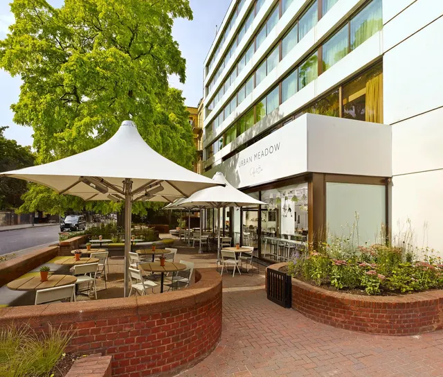 An outdoor dining terrace with white umbrellas, wooden tables and chairs, and brick planters beside a cafe frontage on a hotel building.