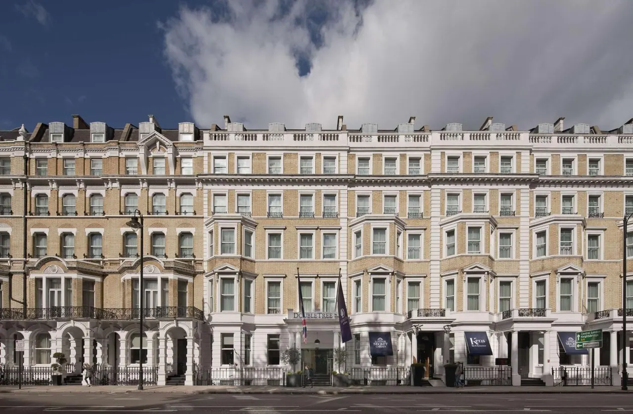 Exterior of a multi-storey cream-coloured Victorian-style hotel with a DoubleTree sign above the entrance, seen from the street under a partly cloudy sky.