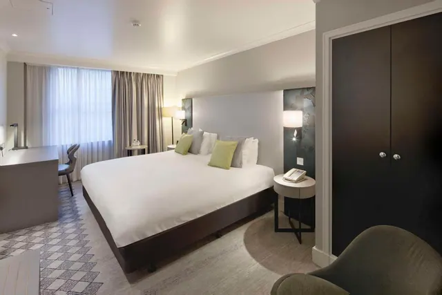 A king bed with white bedding and green cushions sits beside a work desk, a wardrobe and a bedside table with a telephone.