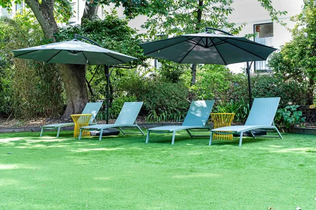 Four sun loungers and two cantilever parasols stand on artificial grass in a garden, with trees and a white building behind.