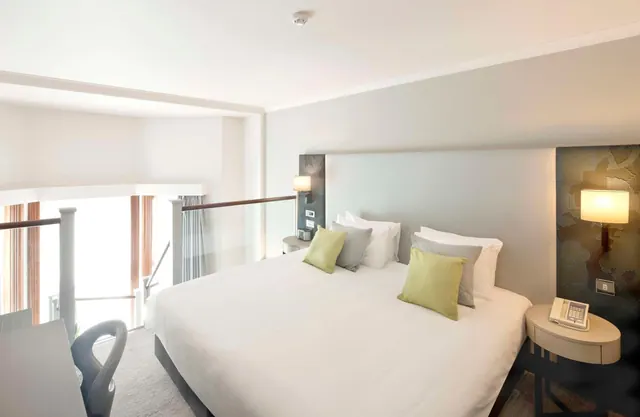 A large white bed with green and grey cushions sits beside a glass railing, with bedside lamps and a telephone on a round side table.