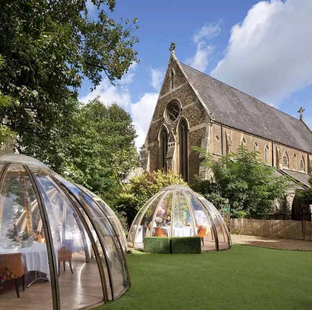 Two transparent dome dining pods stand on a lawn beside a brick Gothic church, with trees and a wooden fence nearby.