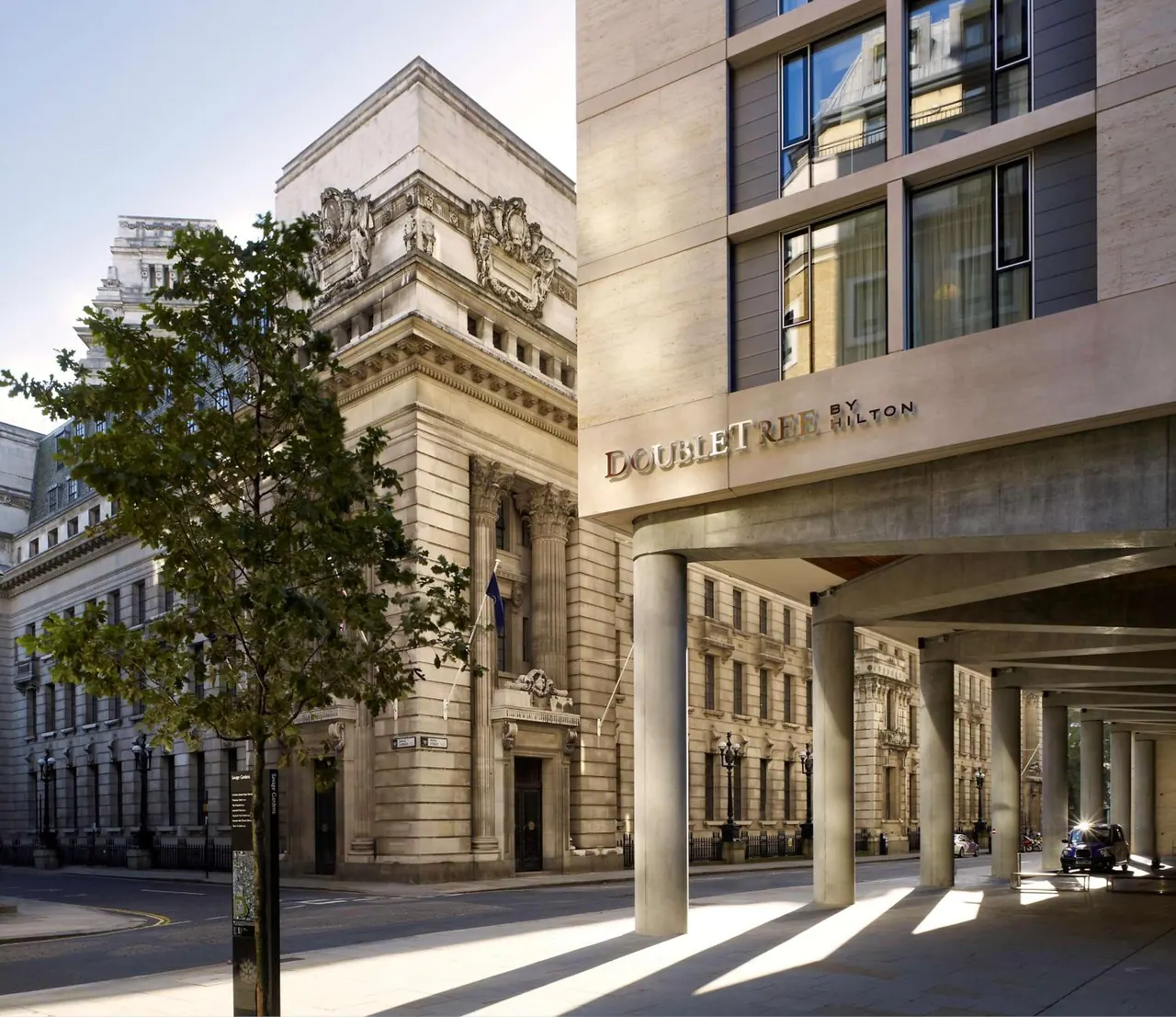 Exterior of a DoubleTree by Hilton building with a covered entrance and concrete columns beside an ornate stone heritage building on a sunlit street.