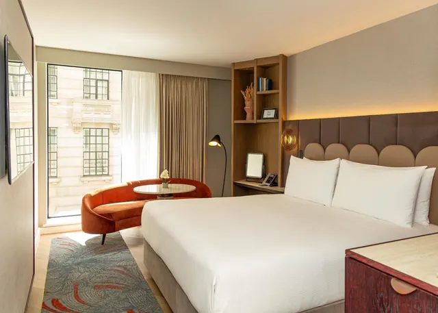 A hotel bedroom with a king bed, a curved orange seating area by a tall window, a floor lamp, and wooden shelving on the wall.