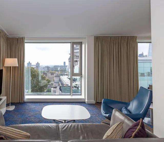 A hotel living area with a grey sofa, a blue armchair and a white coffee table, with large windows looking out over Tower Bridge and the city beyond.